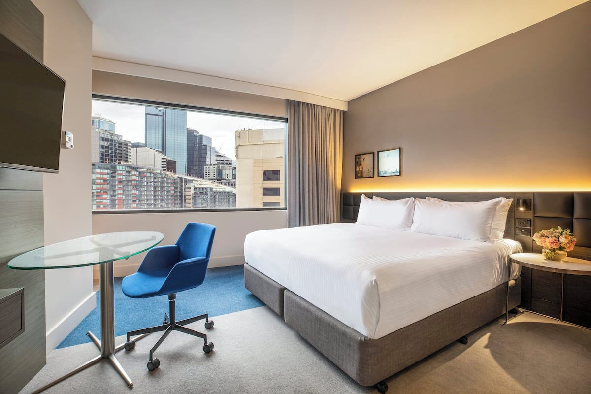 A hotel room with a large bed, a glass table, a blue chair, a nightstand with a vase of flowers, and a view of a cityscape through a large window.