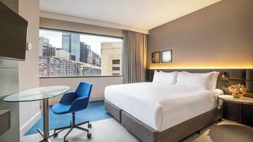 A hotel room with a large bed, a glass table, a blue chair, a nightstand with a vase of flowers, and a view of a cityscape through a large window.
