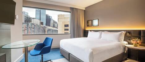 A hotel room with a large bed, a glass table, a blue chair, a nightstand with a vase of flowers, and a view of a cityscape through a large window.