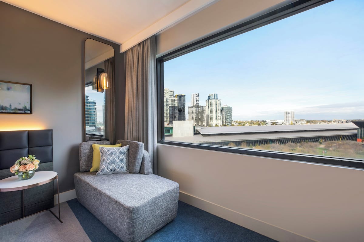 A modern hotel room with a large window, a grey sofa with cushions, a small round table with a vase of flowers, and a cityscape view.