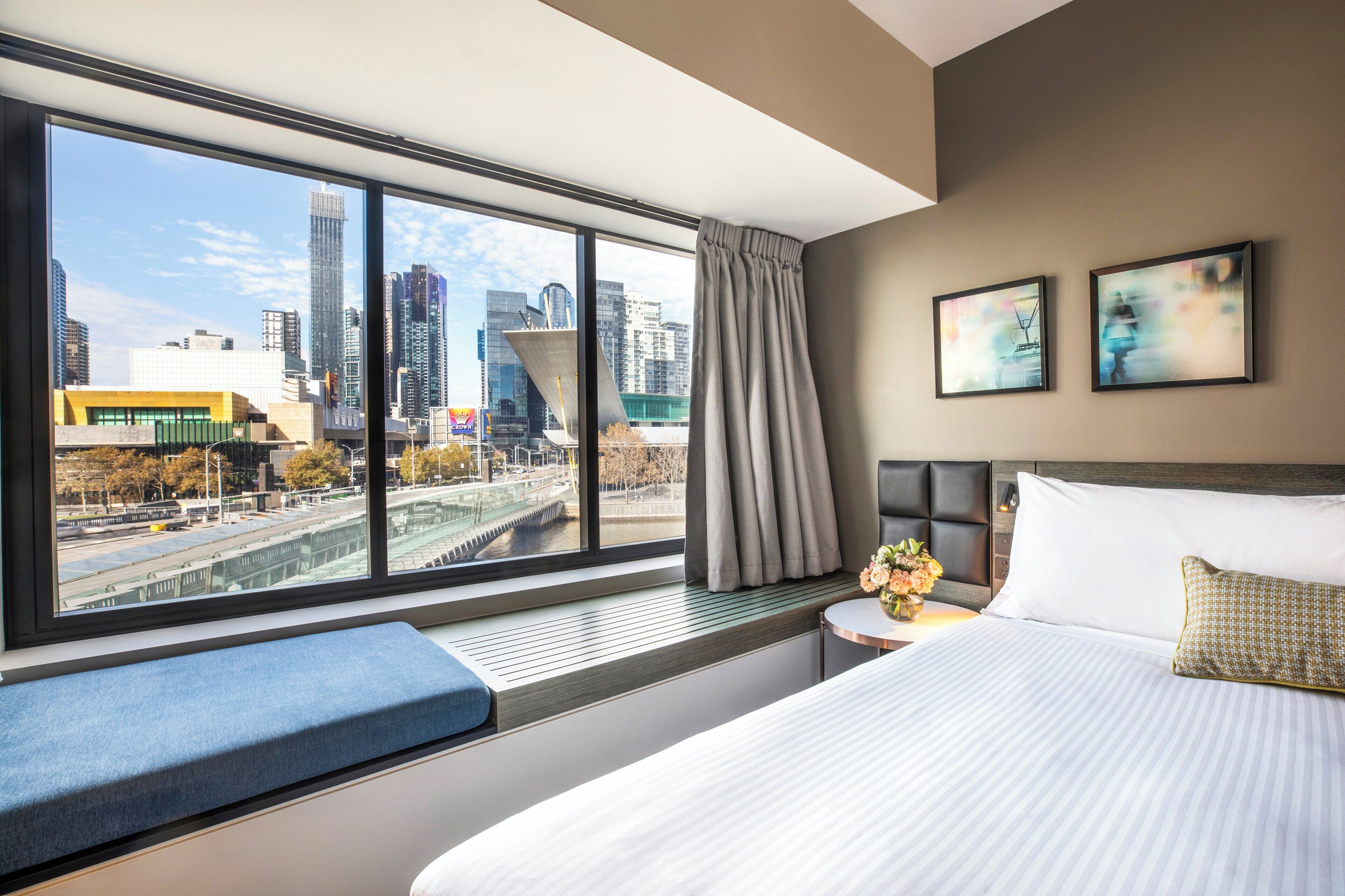 A modern hotel room with a large window offering a city view, a neatly made bed, a bedside table with a vase of flowers, and a bench by the window.