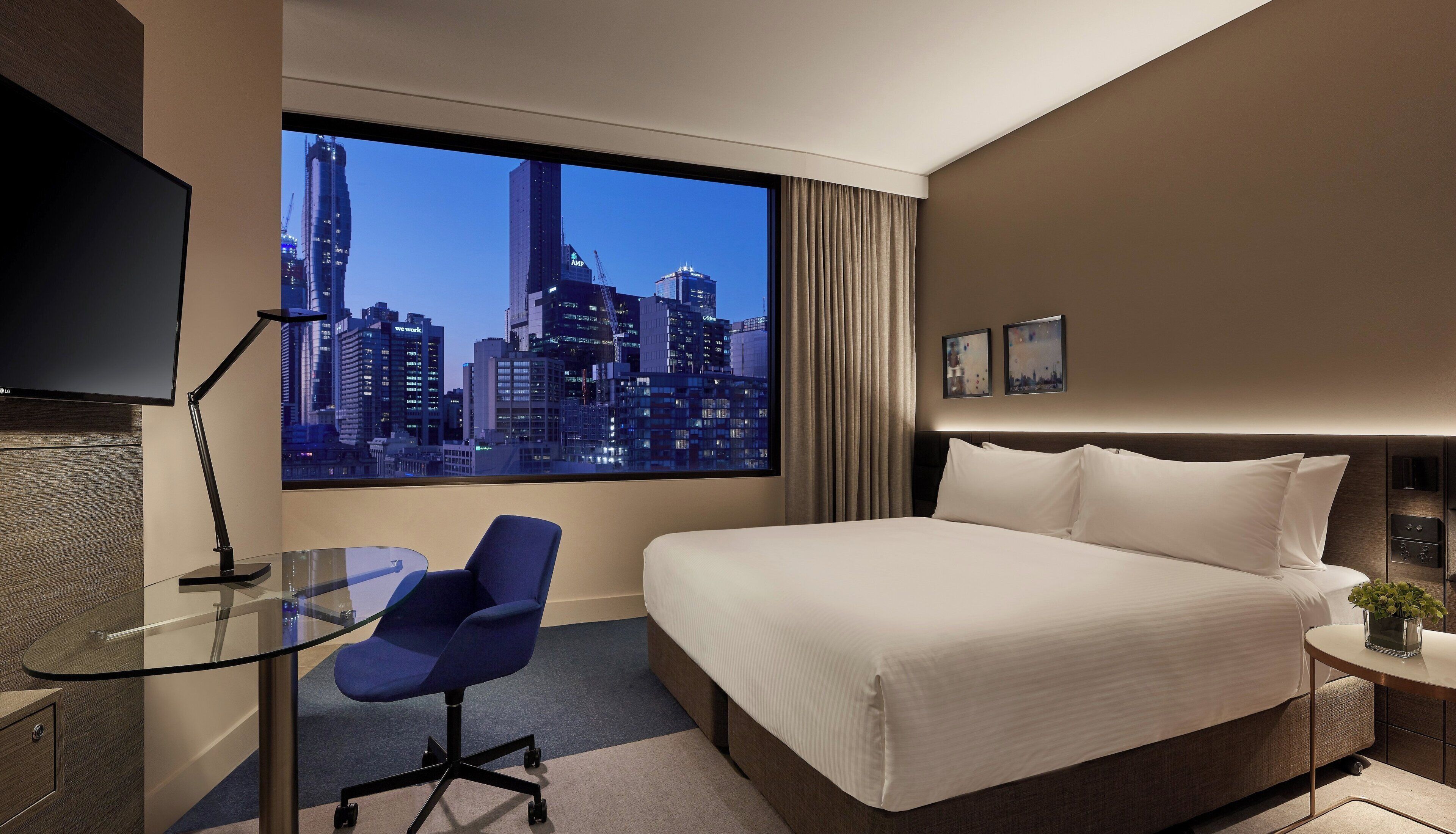 A hotel room with a large bed, a desk with a chair, a TV, and a cityscape view.
