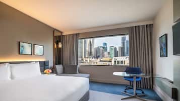 A hotel room with a large bed, a desk with a chair, a view of the cityscape, and a city skyline visible through the window.