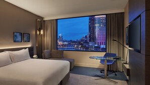 A hotel room with a large bed, a seating area, a desk, and a view of a cityscape at dusk. - Crowne Plaza Melbourne by IHG (Docklands)
