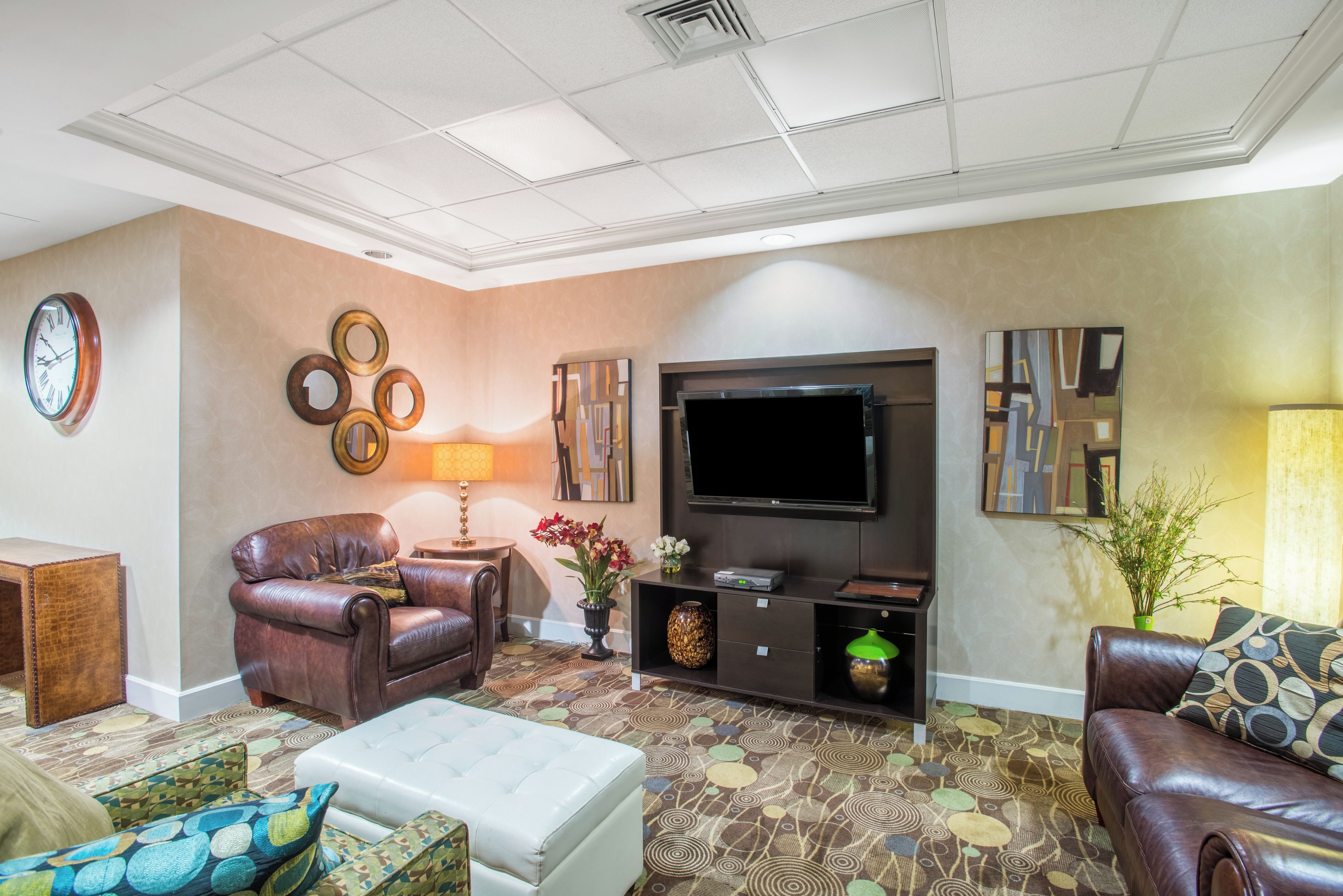 A living room with a leather sofa, a white ottoman, a patterned rug, a wall clock, and a flat-screen TV.