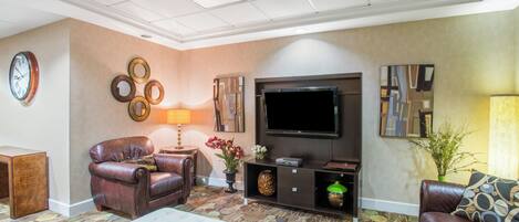 A living room with a leather sofa, a white ottoman, a patterned rug, a wall clock, and a flat-screen TV.