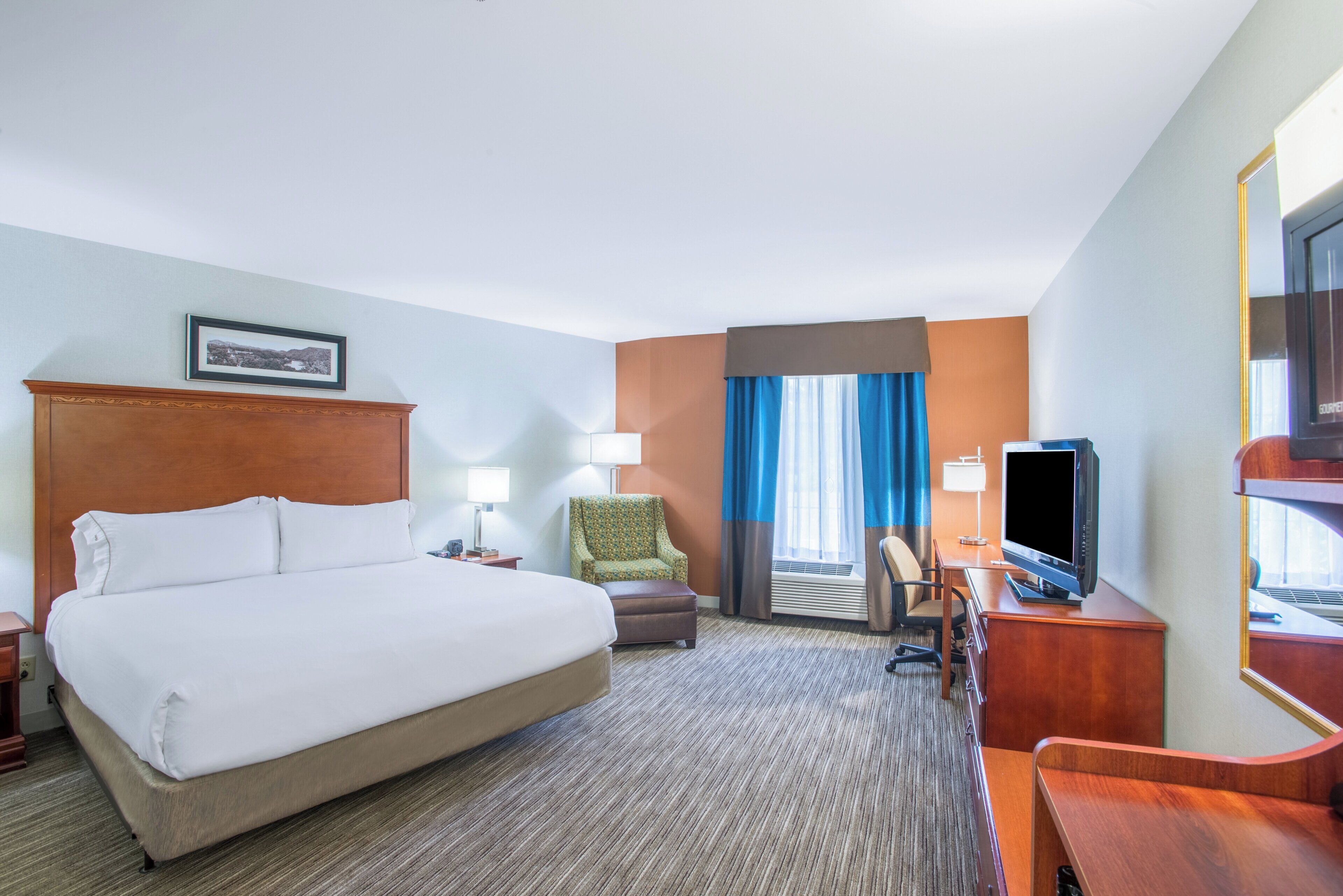 Habitación de hotel con una cama grande, un escritorio con televisor, una silla y una ventana con cortinas.