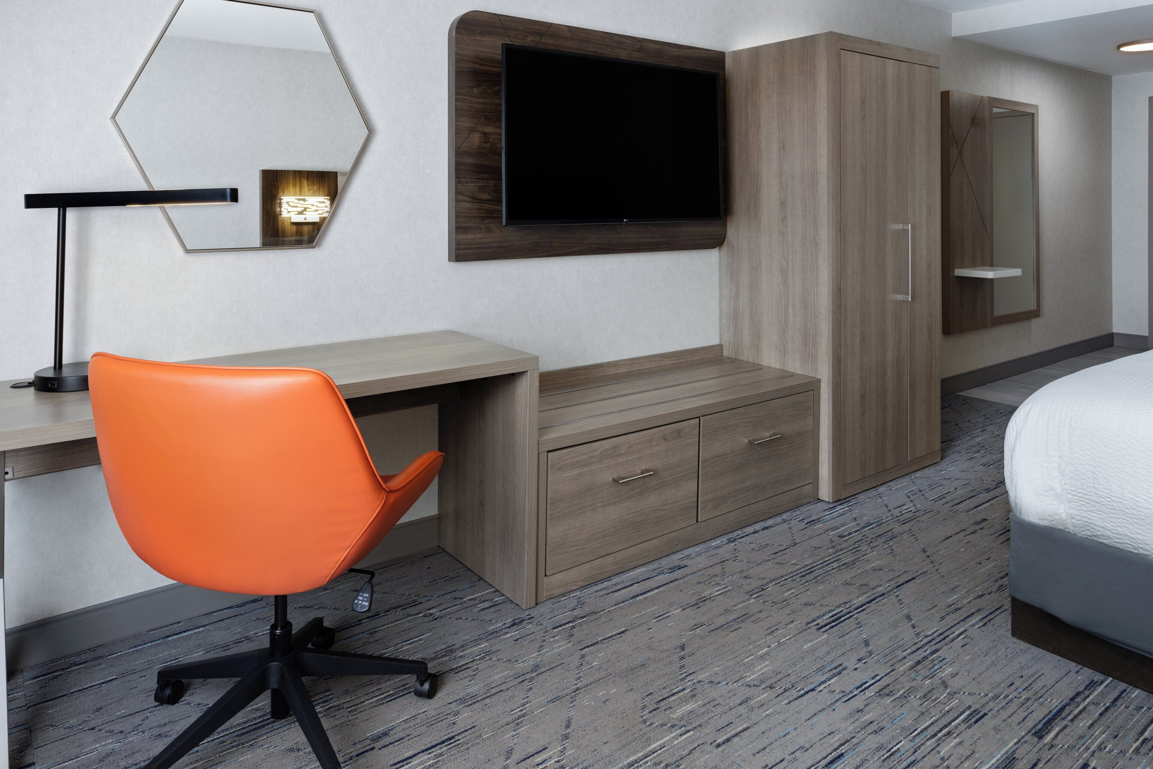 A hotel room with a desk, an orange chair, a television, a wardrobe, and a bed.