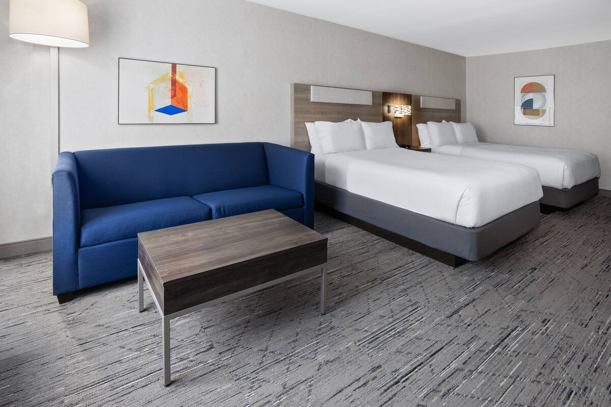 A hotel room with a blue sofa, two single beds, a wooden coffee table, and abstract art on the walls.
