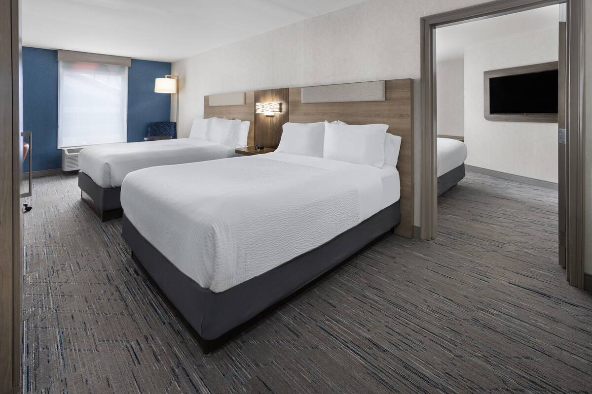 A hotel room with two beds, a flat-screen TV mounted on the wall, a bedside table with a lamp, and a large window with a view.