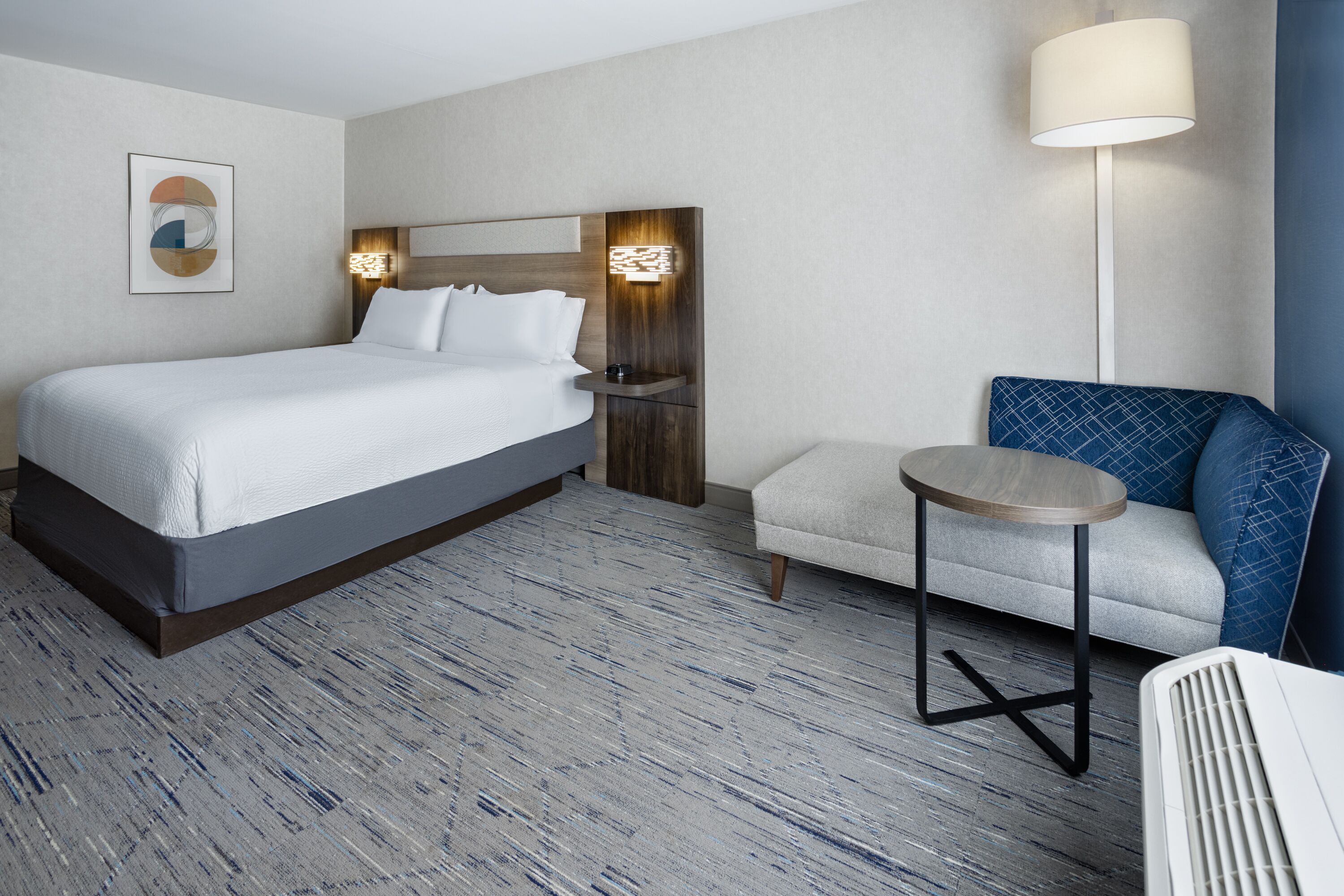 A hotel room with a large bed, a small round table, a sofa, and a standing lamp.