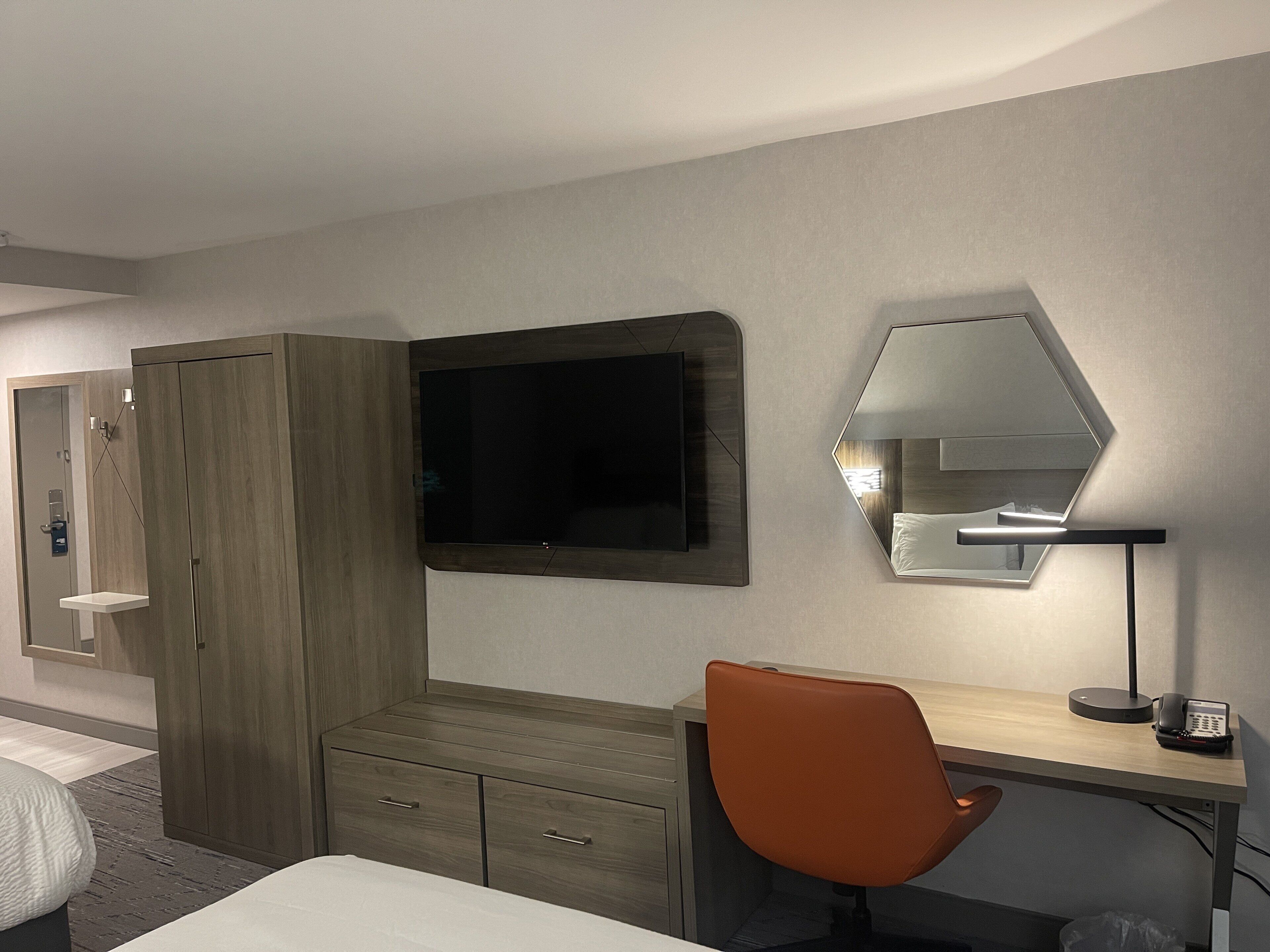 A hotel room with a bed, a desk with a chair, a TV, a mirror, and a wardrobe.
