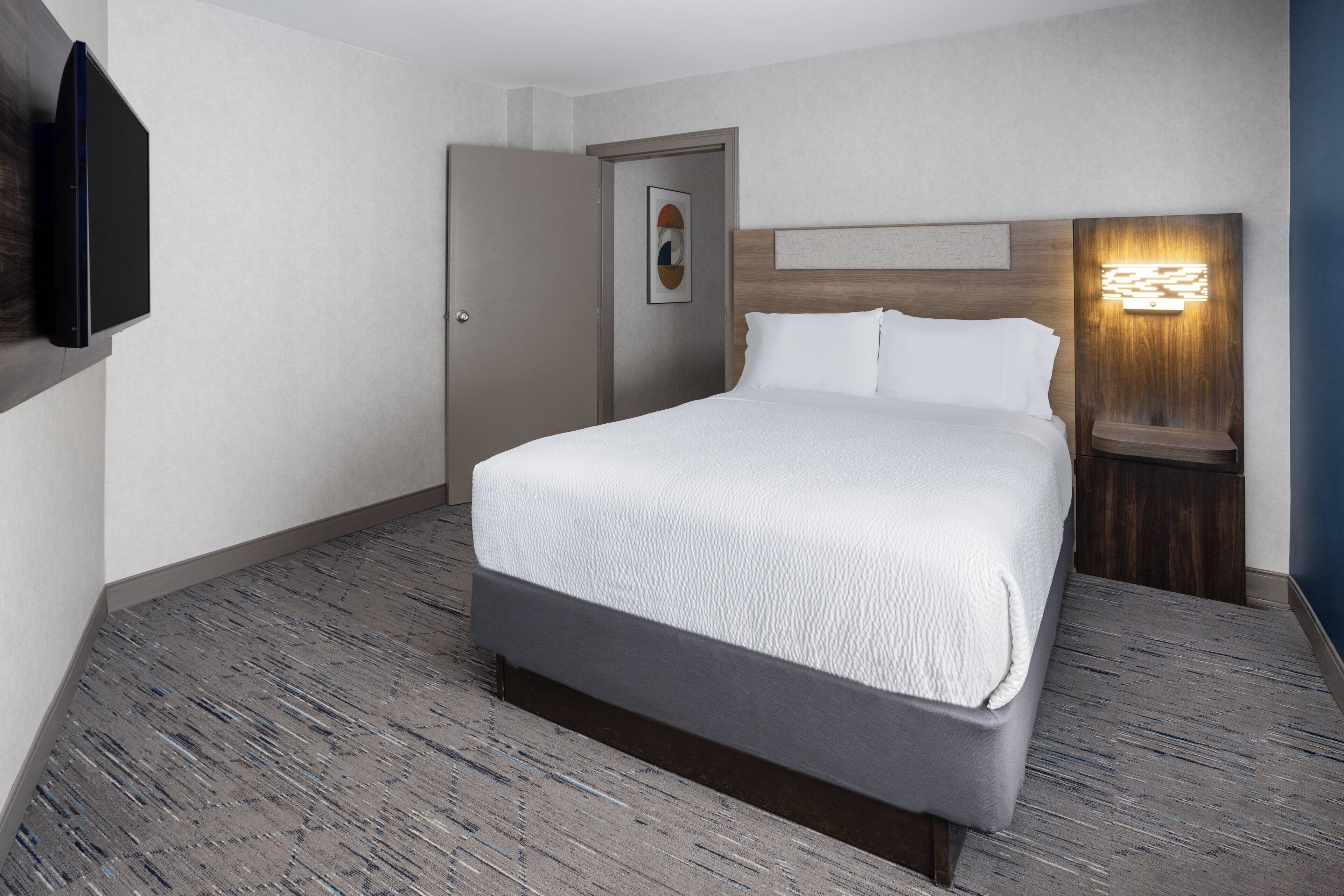 A hotel room with a bed, a door, a wall-mounted TV, and a nightstand with a lamp.