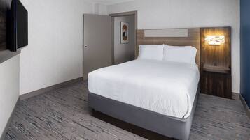 A hotel room with a bed, a door, a wall-mounted TV, and a nightstand with a lamp.