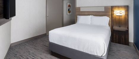 Una habitación de hotel con una cama, una puerta, un televisor montado en la pared y una mesita de noche con una lámpara.