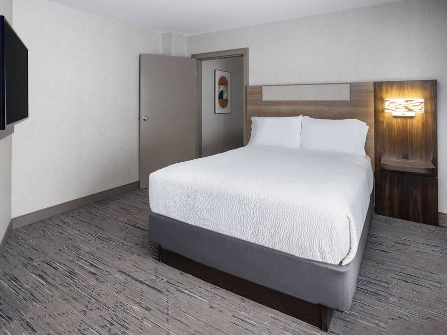 A hotel room with a bed, a door, a wall-mounted TV, and a nightstand with a lamp.