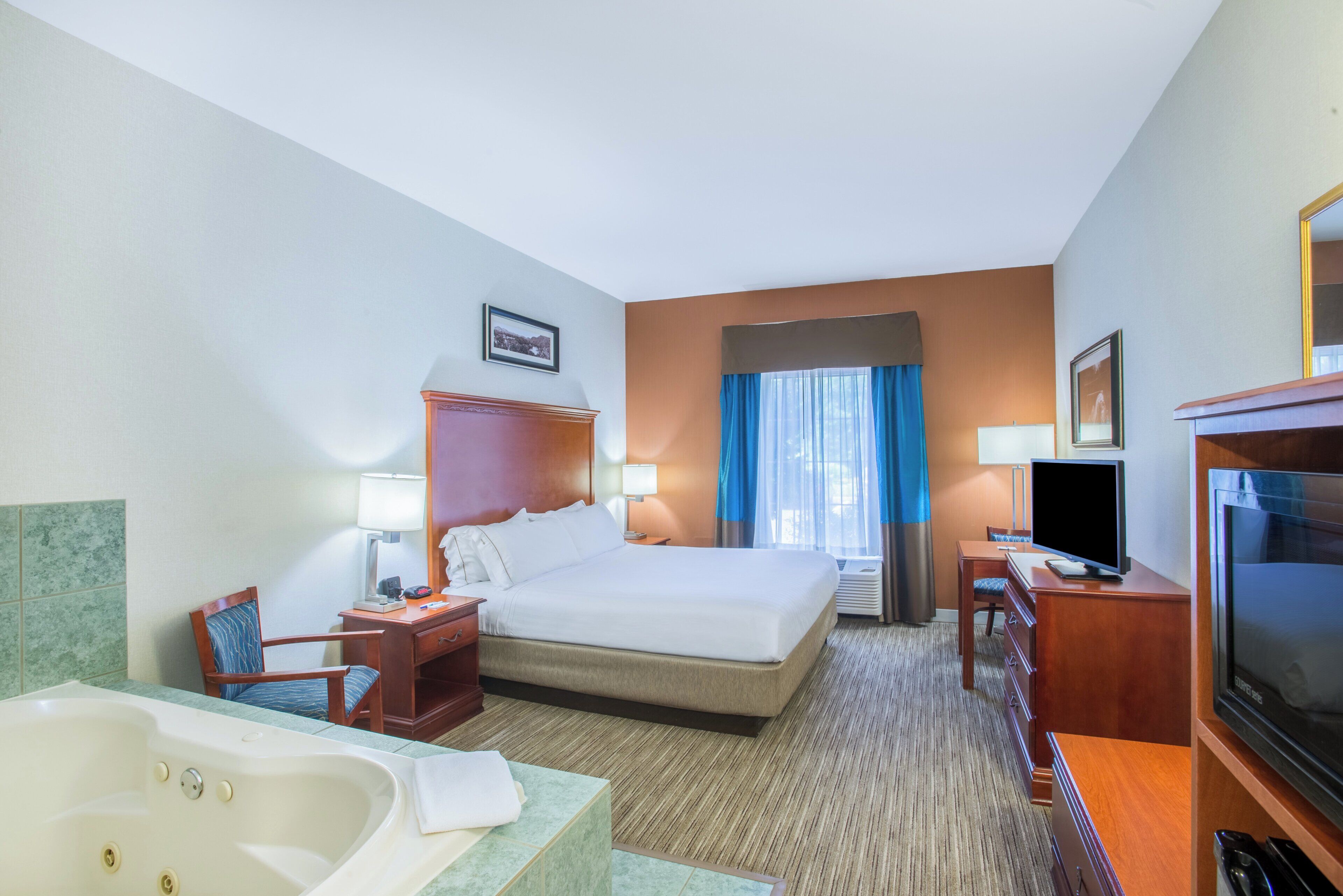 A hotel room with a large bed, a TV, a desk, a chair, a bathroom with a bathtub, and a window with curtains.