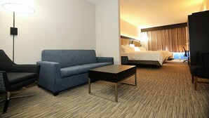 Une chambre d’hôtel avec un canapé bleu, une table basse noire, un lit avec du linge de maison blanc et une grande fenêtre avec des rideaux.