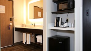 Une salle de bain d’hôtel avec une porte en bois, un lavabo avec un miroir, un porte-serviettes, une machine à café et un réfrigérateur noir.
