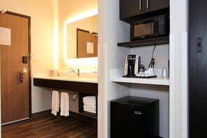 A hotel bathroom with a wooden door, a sink with a mirror, a towel rack, a coffee maker, and a black refrigerator. - Holiday Inn Express & Suites Ashland by IHG (Ashland)