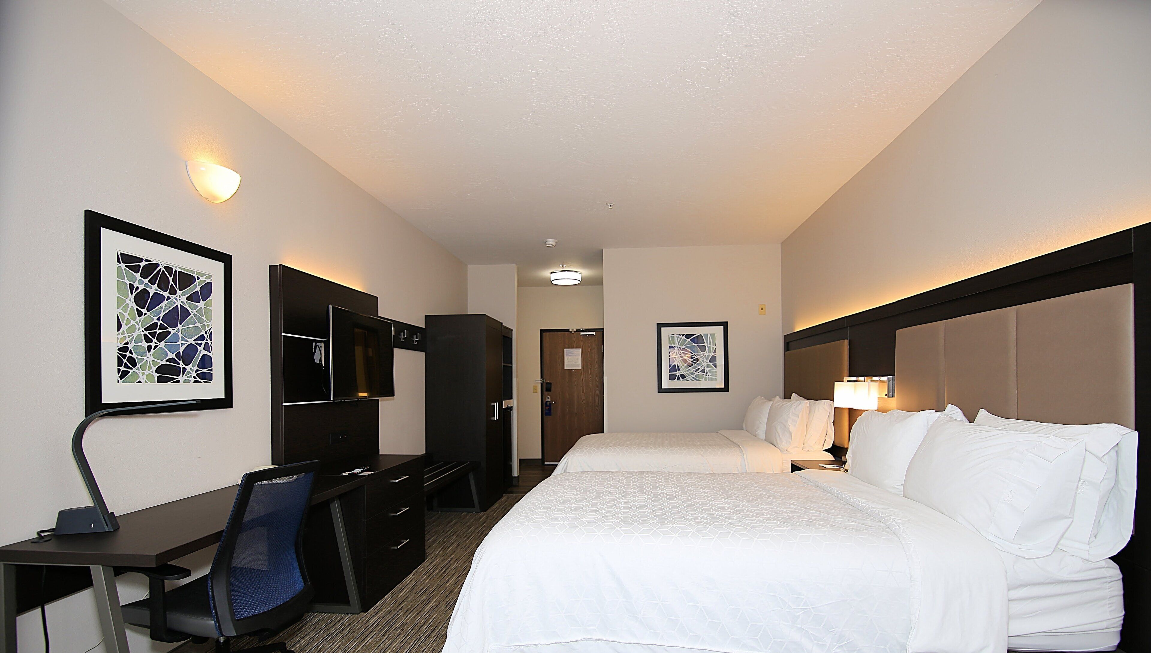 A hotel room with two beds, a desk, a chair, a TV, and artwork on the walls.