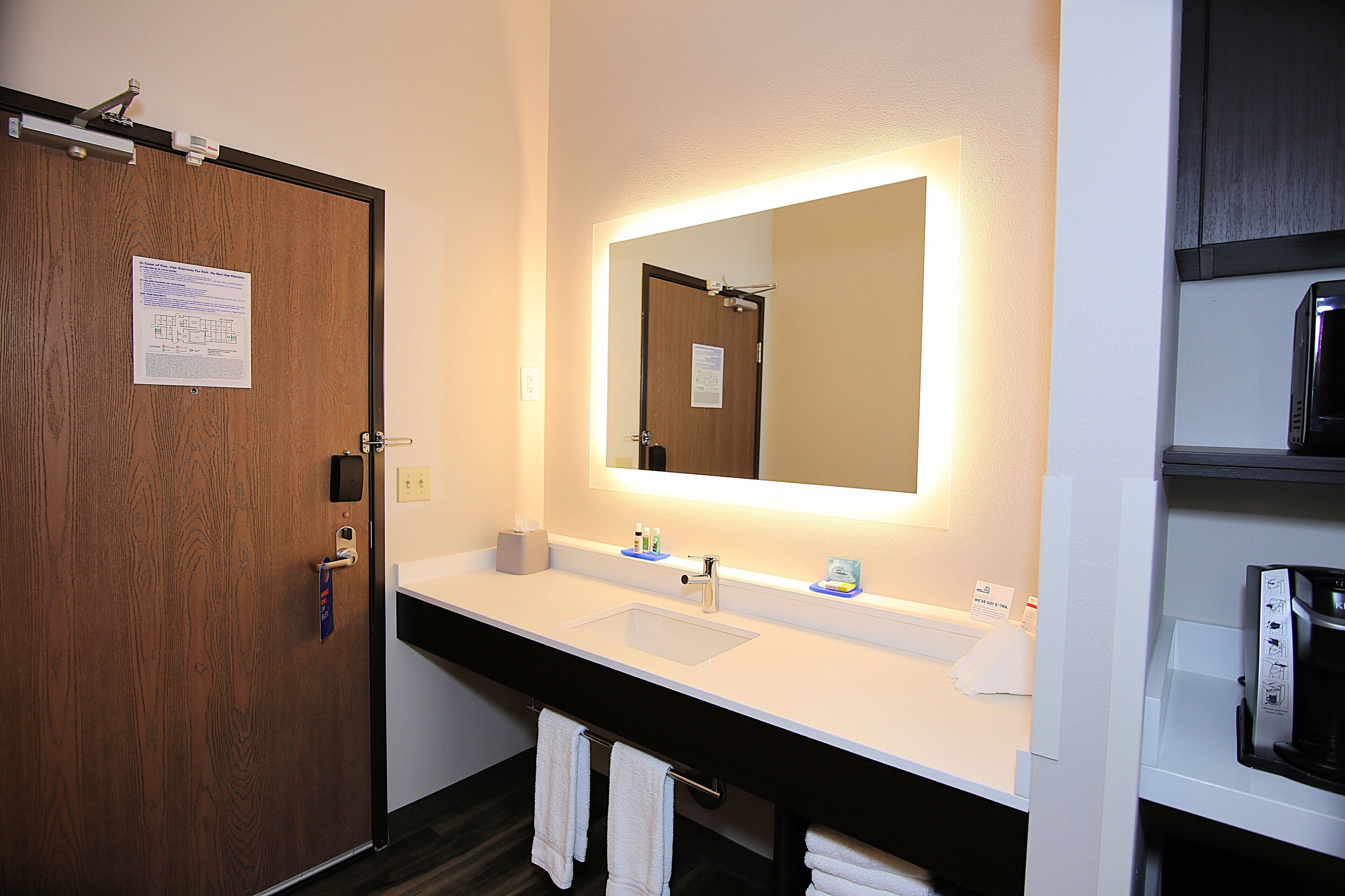 A hotel bathroom with a wooden door, a white sink, a mirror, and a coffee machine.