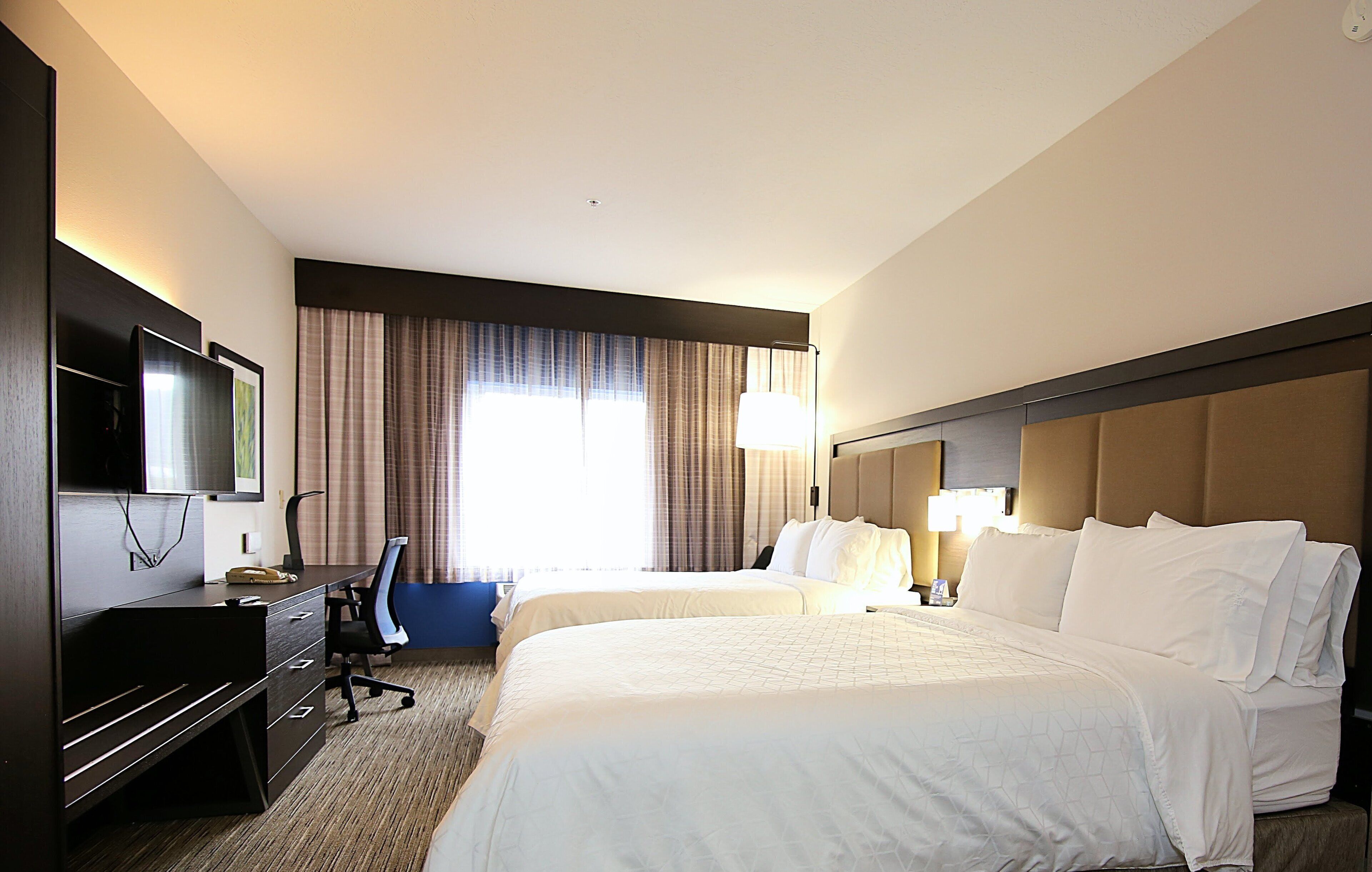 A hotel room with two beds, a desk, a chair, a television, and a large window with curtains.