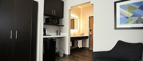 Una habitación de hotel moderna con un armario de madera oscura, un refrigerador negro, un microondas, un lavamanos y una silla.