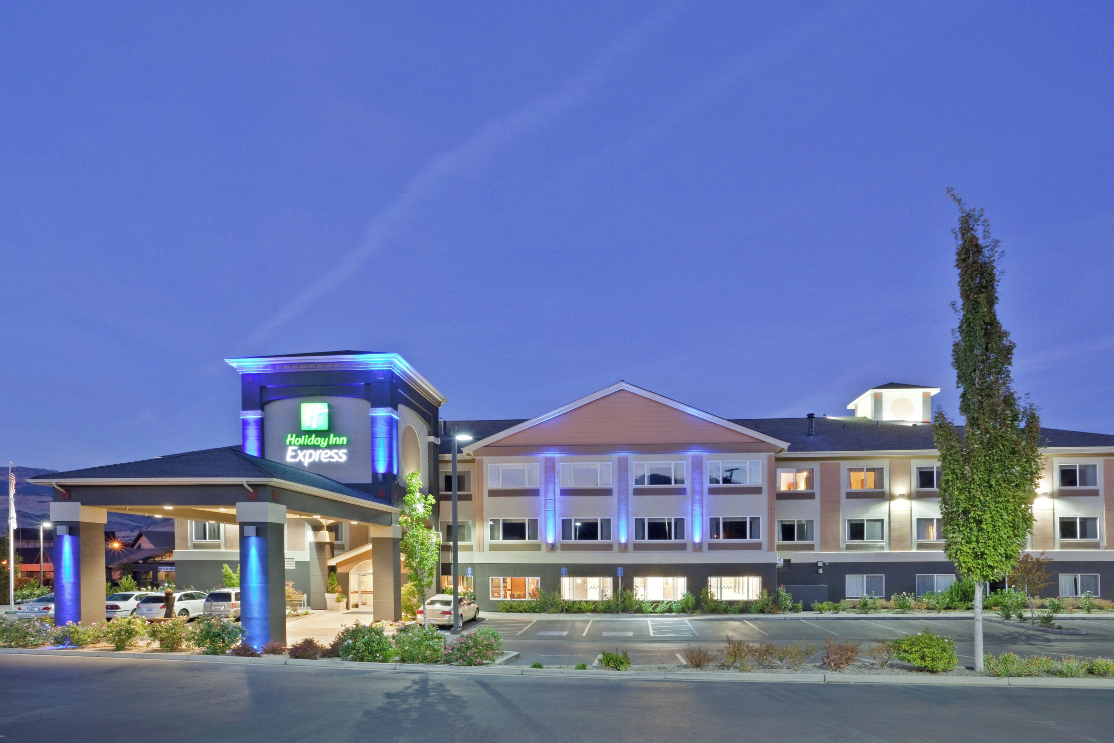 Ein Holiday Inn Express Hotel mit zweistöckiger Bauweise, einem Parkplatz und einer begrünten Fläche mit Sträuchern und einem hohen Baum.