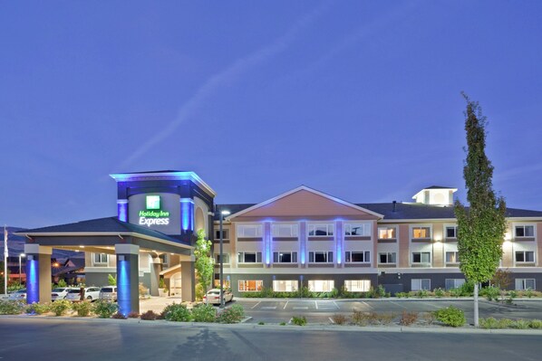 Un hotel Holiday Inn Express con una estructura de dos pisos, un estacionamiento y un área ajardinada con arbustos y un árbol alto.