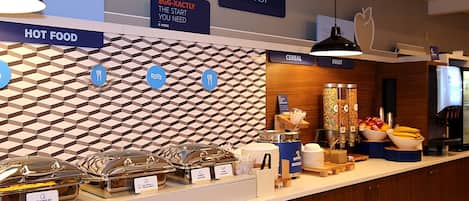 A breakfast buffet area with labeled food stations, including hot food, cereal, and fruit.