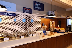 Zona de desayuno tipo buffet con estaciones de comida identificadas, que incluyen alimentos calientes, cereales y fruta.