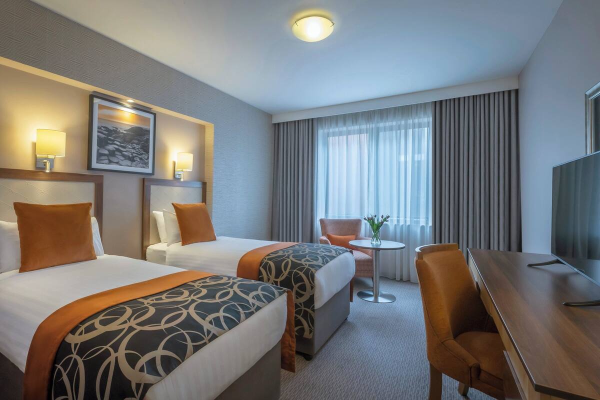 A hotel room with two beds, a desk, a chair, a TV, and a window with curtains.
