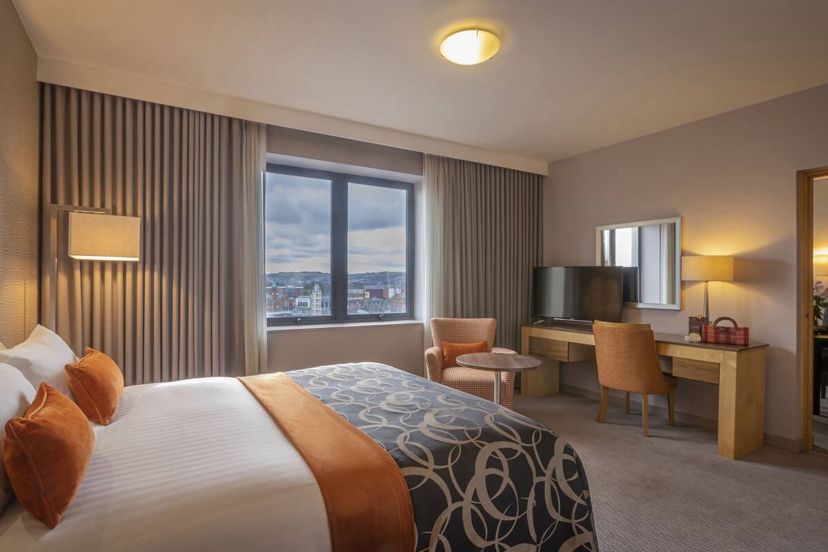 A hotel room with a large bed, a desk with a TV, a chair, and a view of the city through the window.