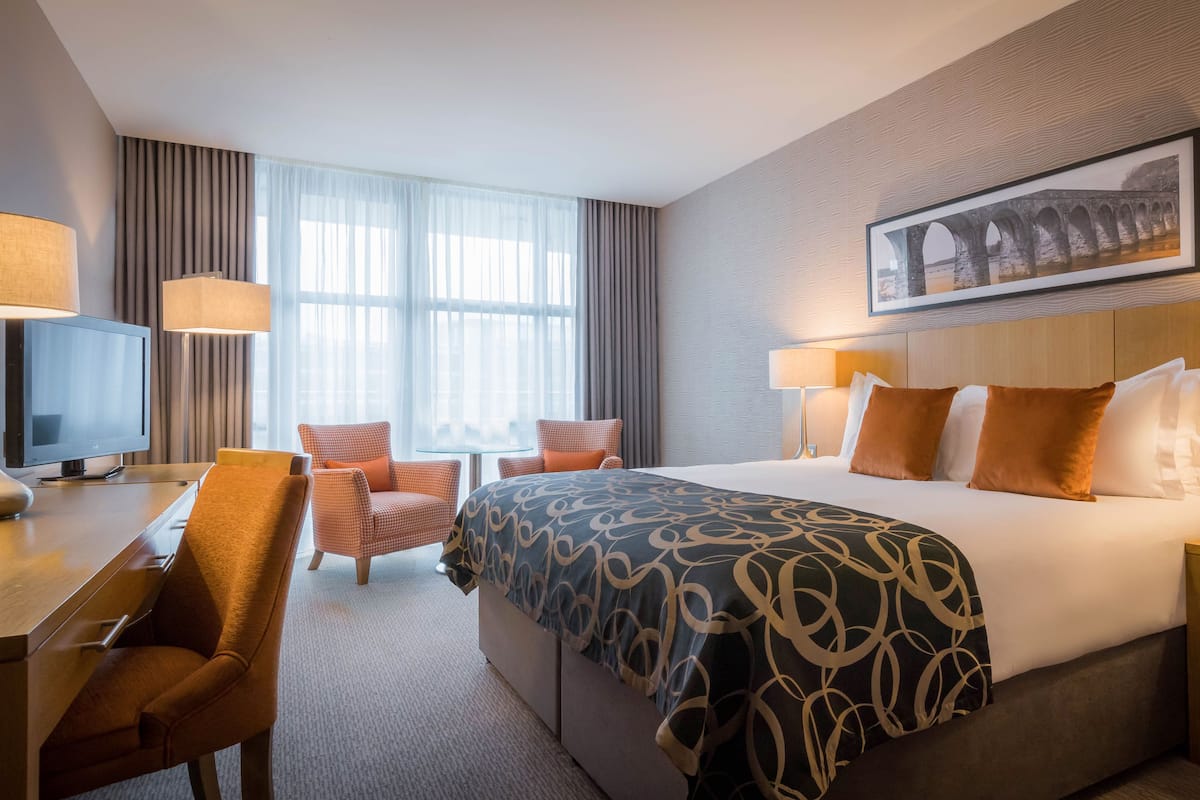 A hotel room with a large bed, two chairs, a desk, a television, and a framed picture on the wall.