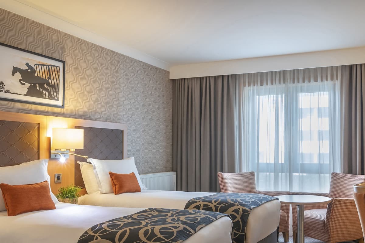 A hotel room with two beds, a small round table, and a chair. There is a large window with curtains, a wall-mounted lamp, and a framed picture on the wall.