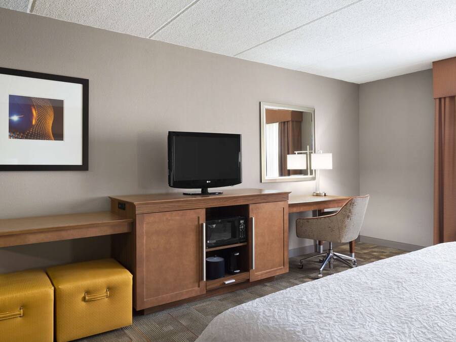 Habitación de hotel con una cama, un televisor en un mueble de madera, un microondas, una silla y un cuadro en la pared.