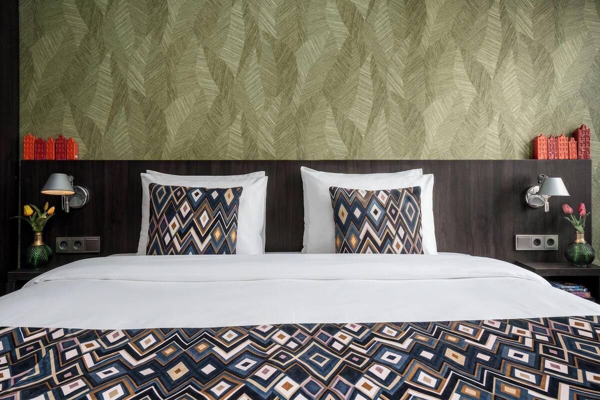 Habitación de hotel con cama, almohadas estampadas, mesita de noche con lámpara y pared con diseño geométrico.