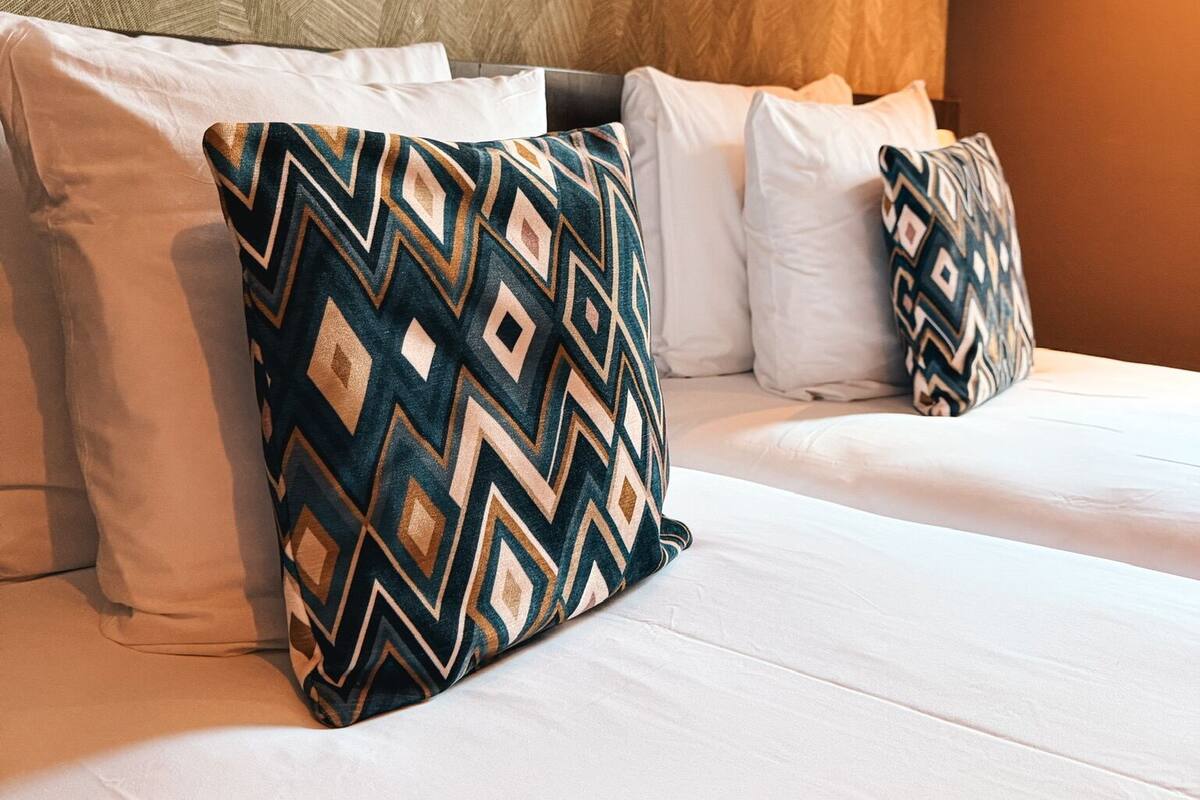 Una cama con sábanas blancas y almohadas estampadas, una pared con textura al fondo y una pared lisa a la derecha.