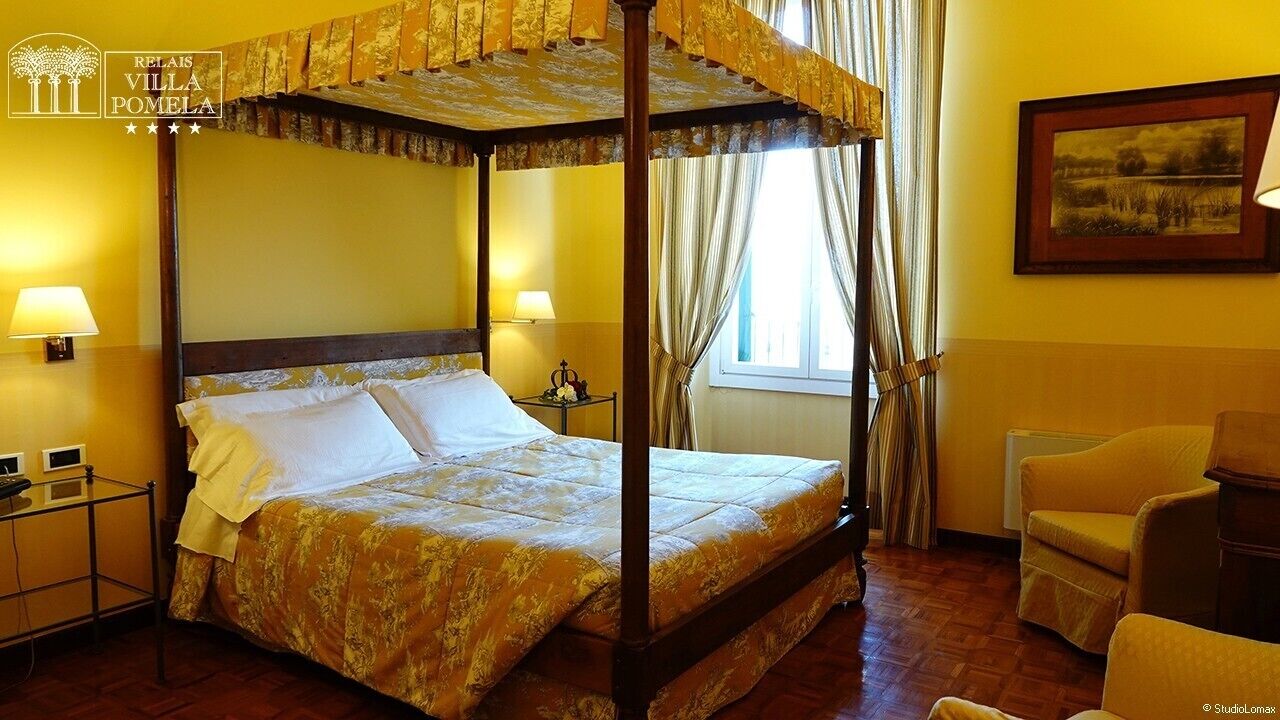 A four-poster bed with a canopy, two bedside lamps, a framed landscape painting, and a chair in a warmly lit room with yellow walls.