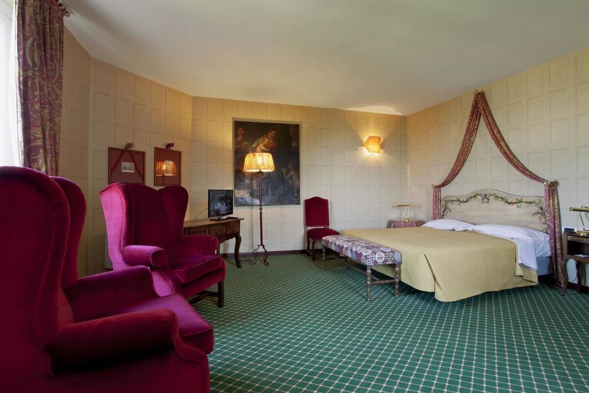 A hotel room with a four-poster bed, a desk with a lamp, and two red armchairs.