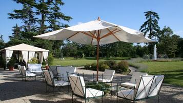 An outdoor seating area with white chairs and a large umbrella, surrounded by trees and a fountain.