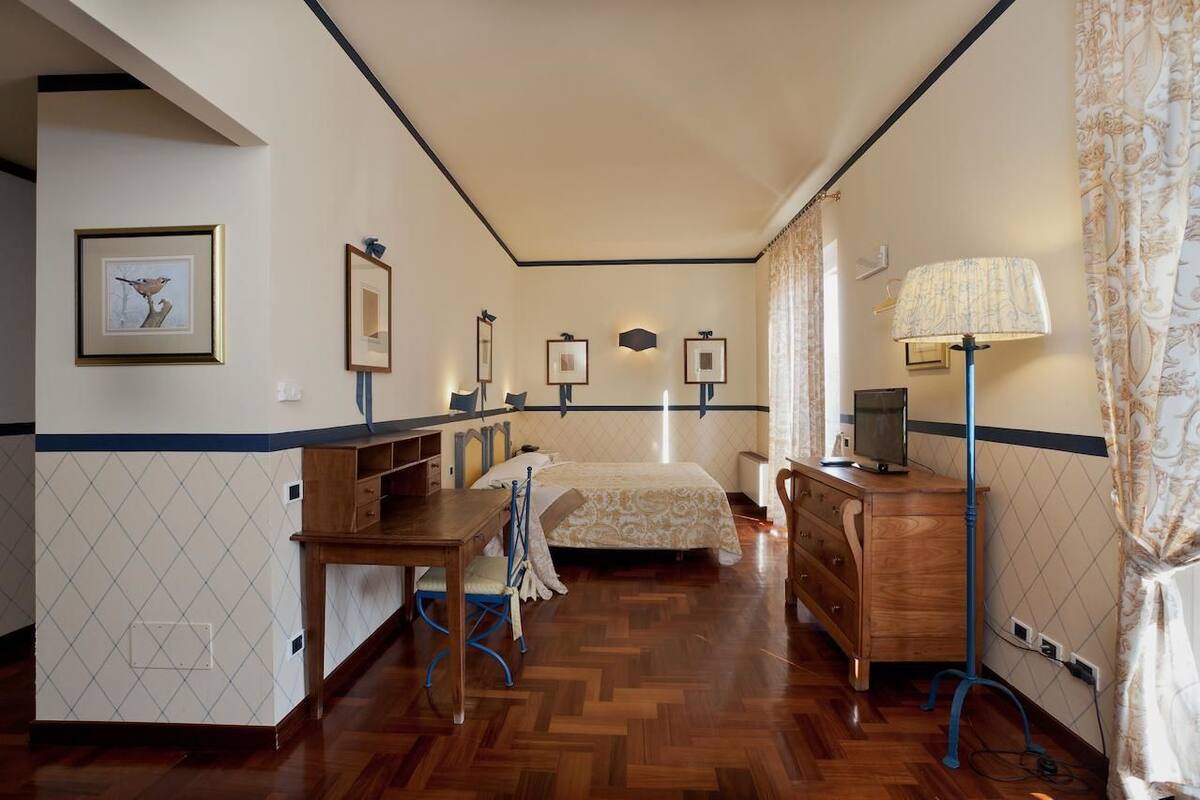 A hotel room with a wooden floor, a bed with a patterned bedspread, a desk with a chair, a dresser, and a lamp.