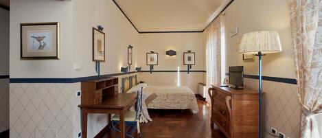 A hotel room with a wooden floor, a bed with a patterned bedspread, a desk with a chair, a dresser, and a lamp.