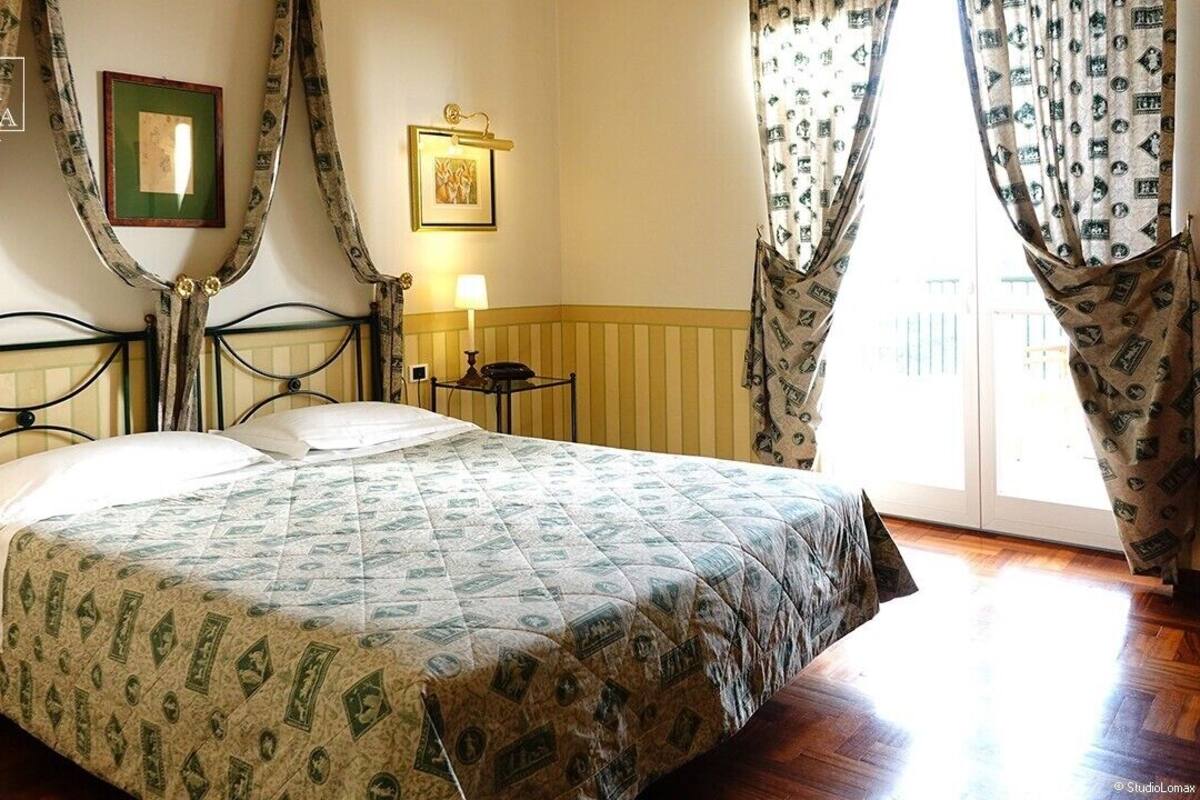A hotel room with a bed, two bedside tables, a nightstand, a lamp, a framed picture on the wall, and curtains.
