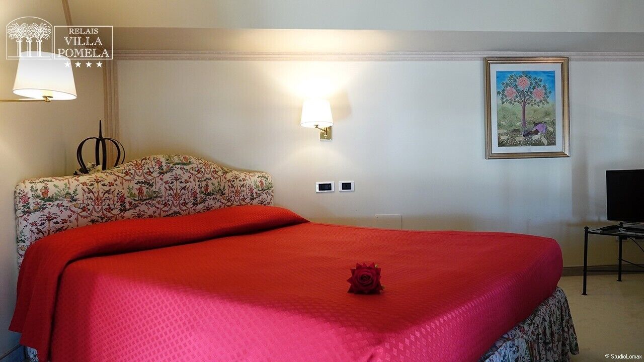 Habitación de hotel con una cama grande, cabecera con estampado floral, colcha roja, una sola rosa como adorno, lámpara de pared, un cuadro enmarcado y un televisor sobre un pedestal.