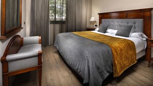 A hotel room with a large bed, a wooden headboard, a grey upholstered chair, and a window with curtains. - Aparthotel Hispanos 7 Suiza (Barcelona)