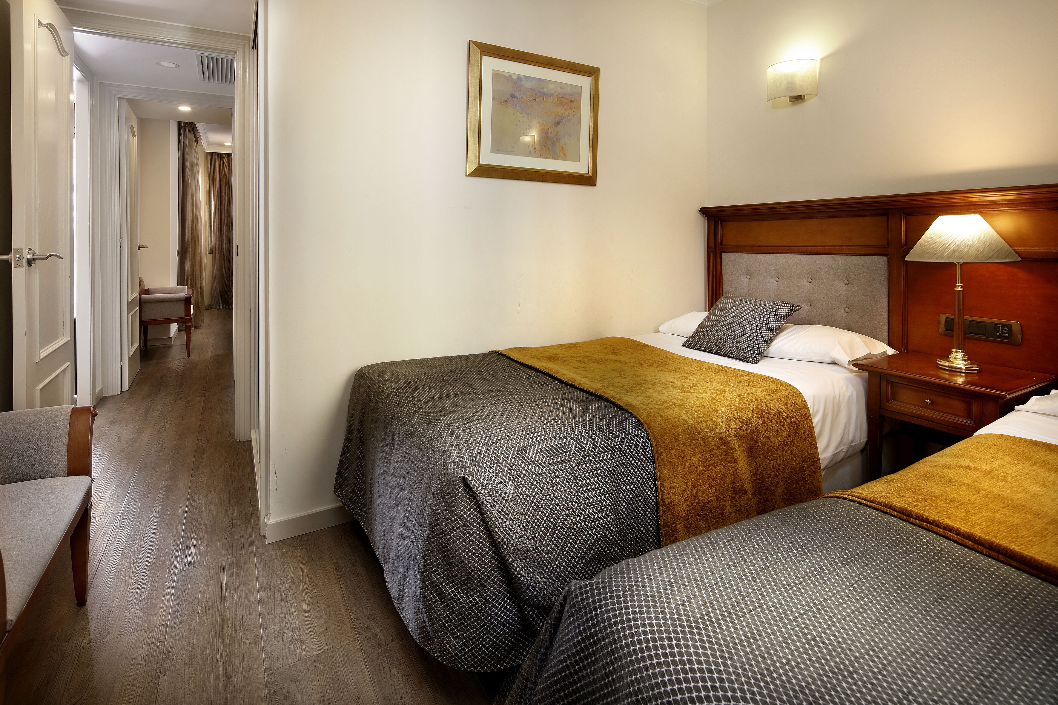 Habitación de hotel con dos camas, cabecero de madera, mesita de noche con lámpara y un cuadro en la pared.