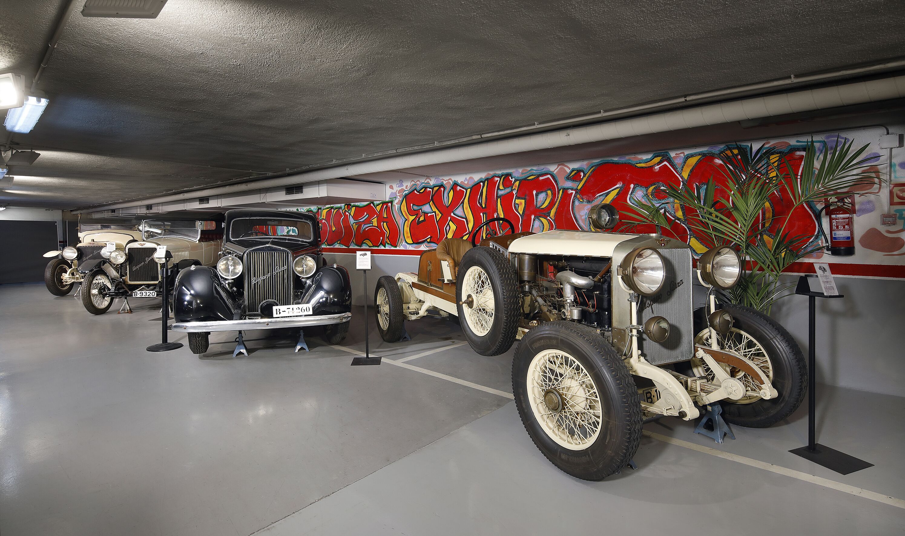 Uma fileira de carros antigos expostos em um museu, com um muro de grafite ao fundo.