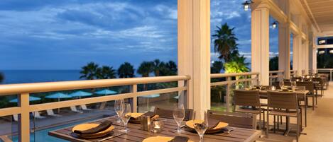 Un espace repas avec des tables dressées pour le dîner, donnant sur une piscine et des palmiers.